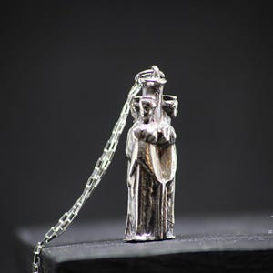 May include: Silver pendant necklace featuring a three-dimensional sculpture of three figures standing together. The figures are wearing robes and appear to be holding hands.