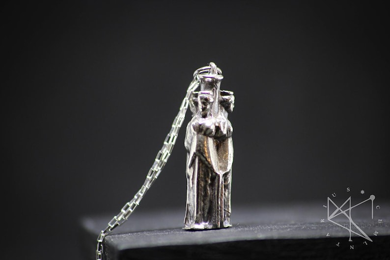 May include: Silver pendant necklace featuring a three-dimensional sculpture of three figures standing together. The figures are wearing robes and appear to be holding hands.
