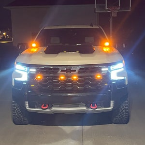 May include: A white Chevrolet pickup truck with a black grille and red tow hooks. The truck has amber lights on the front bumper and side mirrors. The headlights are on and the truck is parked on a paved surface.