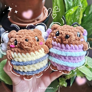 Può includere: Due peluche di api all'uncinetto, uno con pelliccia marrone e strisce gialle e blu, l'altro con strisce viola e blu. Entrambi hanno occhi neri e guance rosa.