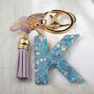 Könnte beinhalten: Ein Schlüsselanhänger mit einem blauen Glitzerbuchstaben K, einem lila Quaste und einem Schmetterlingsanhänger.
