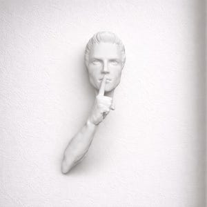 Könnte beinhalten: Eine weiße Wandskulptur, die einen menschlichen Kopf mit einem Finger auf den Lippen darstellt, der Stille signalisiert. Eine Arm erstreckt sich von der Wand, wobei die Hand vor dem Mund positioniert ist. Das Kunstwerk ist an einer strukturierten weißen Wand befestigt.