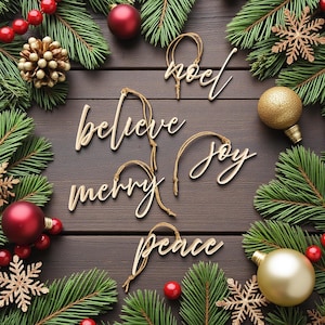 May include: A Christmas-themed composition with wooden word decorations spelling "noel", "believe", "merry", "joy", and "peace". The ornaments are set amongst evergreen boughs, red berries, gold and red baubles, and gold snowflakes on a dark wood surface.