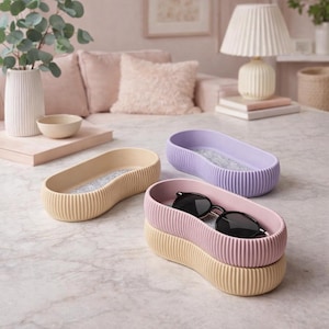May include: Three oval decorative trays in beige, lavender, and pink, with a ribbed exterior and a felt interior. One tray holds black sunglasses. The trays are stacked on a marble surface, with a soft pink sofa in the background.