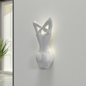 May include: A white wall-mounted sculpture depicting the back of a figure with arms raised above the head. The sculpture has a smooth finish and is illuminated from behind, creating a soft glow.