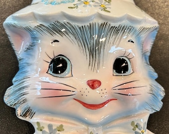 Könnte beinhalten: Ein Wandgefäß aus Keramik in Form eines Katzenkopfes mit einem breitkrempigen Hut. Die Katze hat blaues Fell, große schwarze Augen sowie eine rote Nase und einen roten Mund. Der Hut und eine Schleife unten sind mit kleinen blauen Blumen verziert.