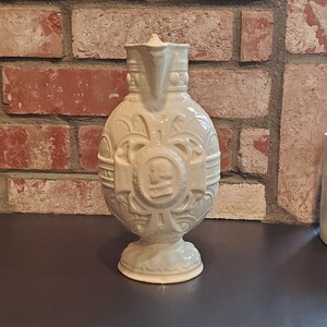 May include: A white ceramic jug with intricate embossed designs, including a central medallion with a face. The jug has a rounded body and a pedestal base.