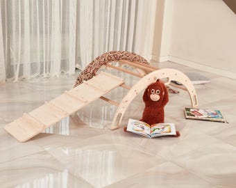 May include: A light-colored wooden climbing arch with a detachable ramp, featuring a brown plush monkey toy holding an open book, a patterned snake-shaped cushion draped over the top, and a second book lying flat on the floor nearby.