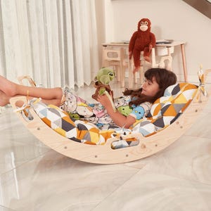 Peut inclure: Un basculeur en bois incurvé avec une finition en bois naturel clair et des poignées découpées sur les côtés. Le basculeur comprend un coussin rembourré avec un motif géométrique dans des tons de jaune, gris, blanc et noir, fixé par des liens.