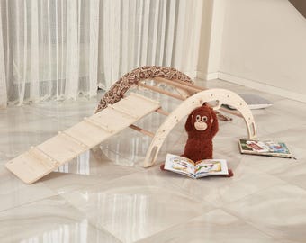 May include: A light-colored wooden climbing arch with a matching ramp attached to one side. A brown plush monkey toy sits in front of the arch, holding an open book with colorful illustrations. A patterned fabric drape is hung over the arch structure.