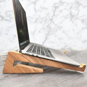 Peut inclure: Un ordinateur portable argenté posé sur un support en bois brun clair au profil triangulaire. Le support présente une découpe et une petite lèvre avant pour maintenir l'appareil en place, offrant une vue inclinée du clavier et de l'écran.