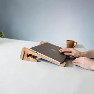 Könnte beinhalten: Ein hellbrauner Laptopständer aus Holz mit geneigtem Design, auf dem ein geschlossener silberner Laptop ruht. Der Ständer besteht aus einer einfachen Steckkonstruktion aus zwei Holzteilen, die dem Gerät einen erhöhten Winkel verleiht.