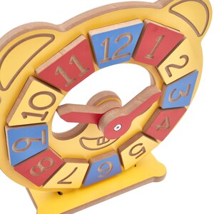 May include: A yellow wooden bear-shaped clock toy featuring twelve removable square blocks numbered one through twelve in red, blue, and yellow. The clock has two red movable hands attached to a central pivot point and a base stand.