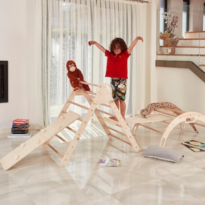 Puede incluir: Un triángulo de escalada de madera con un tobogán, una escalera y una rampa curva. Un niño está escalando el triángulo, y hay un mono de peluche en la parte superior.