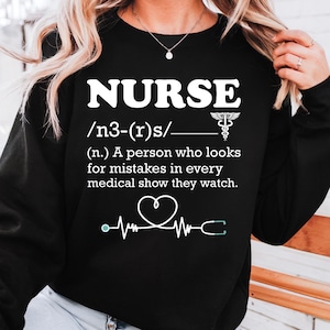 Może przedstawiać: Czarna bluza z napisem "NURSE" w białym kolorze, definicją, symbolem medycznym i grafiką monitora pracy serca. Bluza ma okrągły dekolt.