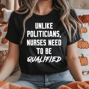 Puede incluir: Camiseta negra con texto blanco: "UNLIKE POLITICIANS, NURSES NEED TO BE QUALIFIED." La camiseta de manga corta se usa con vaqueros azules. El fondo presenta cojines con motivos de calabazas y una cesta de mimbre.