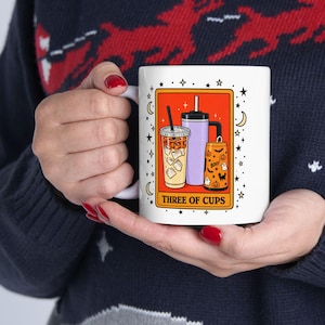 May include: A white ceramic mug featuring a graphic of three different drink containers inside an orange rectangular border. The design includes the text "THREE OF CUPS" and is surrounded by small black stars and crescent moon illustrations.