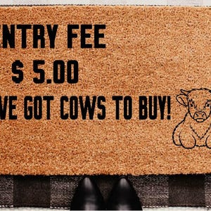 Puede incluir: Una alfombra rectangular con el texto "ENTRY FEE $5.00 I'VE GOT COWS TO BUY!" en negro. Un dibujo lineal de una vaca está a la derecha. La alfombra es marrón y está sobre una alfombra a cuadros en blanco y negro.