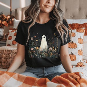 Puede incluir: Camiseta negra con un diseño de fantasma en un campo de flores. El fantasma tiene una cara sencilla. La camiseta es de cuello redondo y manga corta. El diseño está sobre un fondo oscuro. El tema es Halloween y la naturaleza.