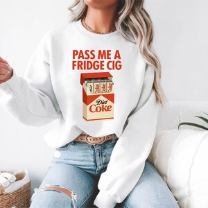 Pu&ograve; includere: Felpa bianca con la scritta "PASS ME A FRIDGE CIG" in lettere rosse. La grafica presenta un pacchetto di sigarette Diet Coke. La felpa &egrave; abbinata a jeans blu.