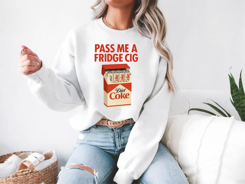 Pu&ograve; includere: Felpa bianca con la scritta "PASS ME A FRIDGE CIG" in lettere rosse. La grafica presenta un pacchetto di sigarette Diet Coke. La felpa &egrave; abbinata a jeans blu.