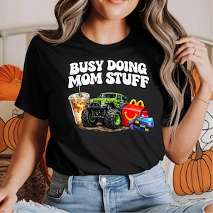 Puede incluir: Camiseta negra con el texto blanco "BUSY DOING MOM STUFF". El diseño incluye un monster truck verde, un café para llevar, una comida rápida y tarjetas de crédito.