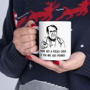 May include: White ceramic mug featuring a black and white illustration of a man pointing. The text on the mug reads, "YOU'RE NOT A PICKY EATER IF YOU ARE 600 POUNDS."