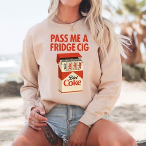 Pu&ograve; includere: Felpa beige con la scritta rossa "PASS ME A FRIDGE CIG". La grafica presenta un pacchetto di Diet Coke con lattine di Diet Coke all'interno. La felpa &egrave; a maniche lunghe e girocollo.