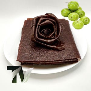 May include: A dark brown fruit leather, shaped like a rose, rests on a stack of flat fruit leather sheets on a white plate. Several green plums are arranged to the side. The fruit leather has a textured appearance.
