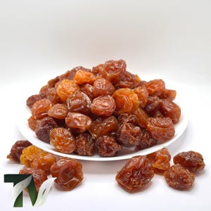 May include: A white plate piled high with dried fruit. The fruit is a mix of amber and dark brown hues, with a slightly wrinkled texture. Some pieces are scattered around the plate, against a white backdrop.