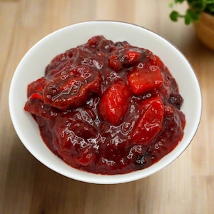 May include: A white bowl filled with a vibrant red cranberry sauce. The sauce has a thick consistency and contains whole cranberries and chunks of fruit. The dish is set on a light-coloured wooden surface.