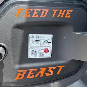 May include: A black gas tank door with an orange sticker that says "FEED THE BEAST".