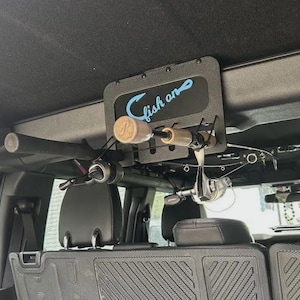 May include: A black metal overhead fishing rod rack mounted to the interior roof of a vehicle. The rack features a rectangular plate with the text "fish on" in light blue script and includes black elastic cords to secure two fishing rods.