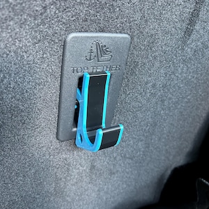 May include: A black plastic car seat top tether anchor with a blue trim. The anchor is labeled "Top Tether" and has a symbol of an anchor and a car seat.