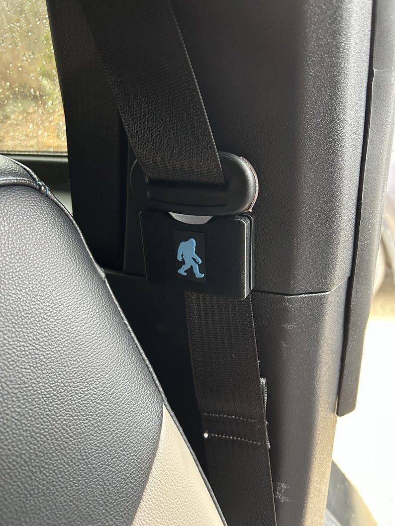 May include: A black car seatbelt with a black buckle and a blue silhouette of a Bigfoot on a black rectangular tag.