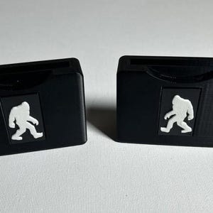 May include: Two black plastic cases with a white silhouette of a Bigfoot walking on each case.