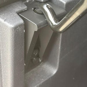 May include: Close-up of a black plastic component with a curved metal handle. The plastic has a textured surface and a series of layered edges. The handle is shiny and reflects light, contrasting with the matte finish of the plastic.