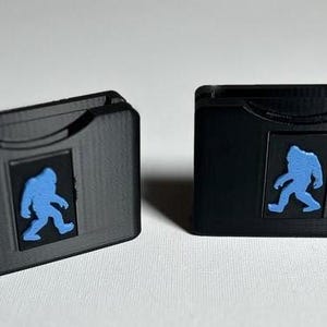 May include: Two black plastic cases with a blue silhouette of a Bigfoot on the front. The cases are designed to hold small items like SD cards or game cartridges.