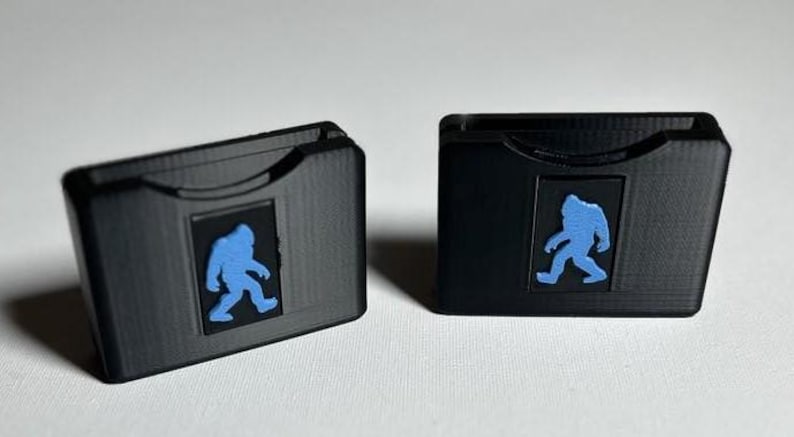 May include: Two black plastic cases with a blue silhouette of a Bigfoot on the front. The cases are designed to hold small items like SD cards or game cartridges.