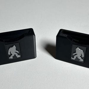 May include: Two black plastic game card holders with a grey silhouette of a Bigfoot walking on each.
