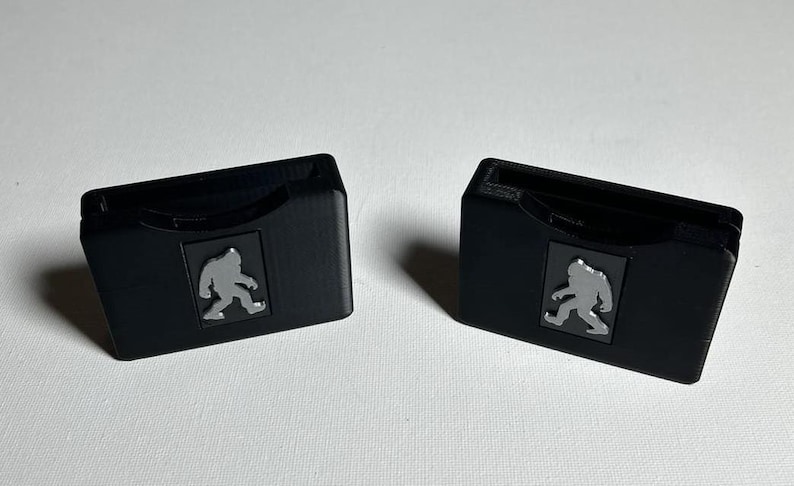 May include: Two black plastic game card holders with a grey silhouette of a Bigfoot walking on each.