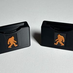 May include: Two black plastic card holders with a cutout for a card. Each holder features a small orange graphic of a Bigfoot walking.