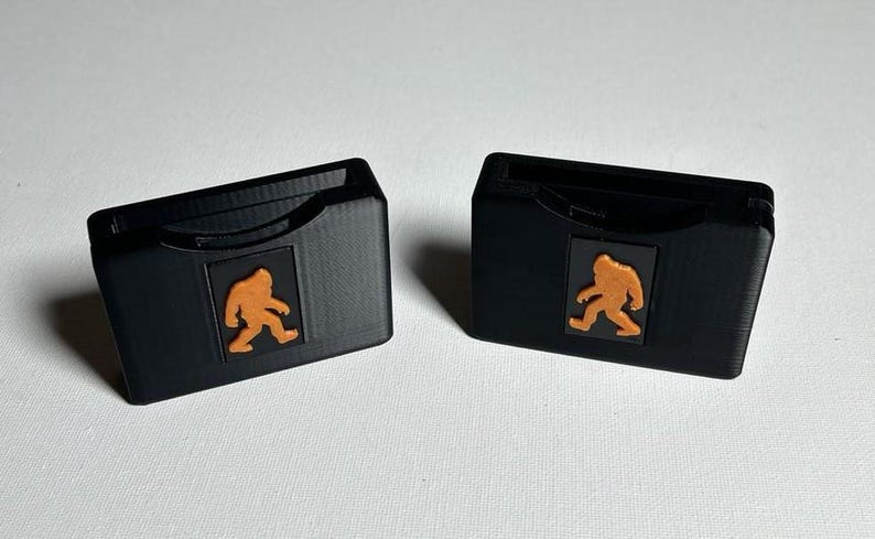 May include: Two black plastic card holders with a cutout for a card. Each holder features a small orange graphic of a Bigfoot walking.