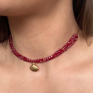 Può includere: Una delicata collana a catena dorata con una fila di perline di cristallo rosso e un ciondolo a forma di conchiglia dorata.