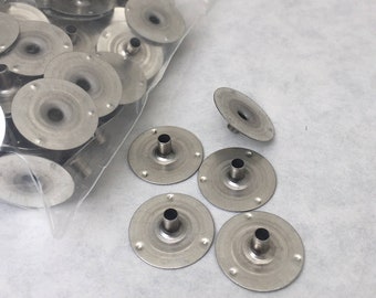 May include: A bag of silver candle holders with a few holders scattered on a white surface. The holders are round with a small hole in the center and a raised lip around the edge.