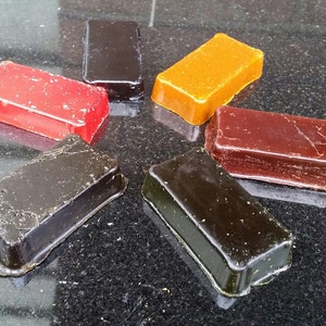 May include: Six rectangular blocks of wax in different colors: red, orange, brown, black, and two shades of dark brown. The blocks are arranged in a star shape on a black surface.