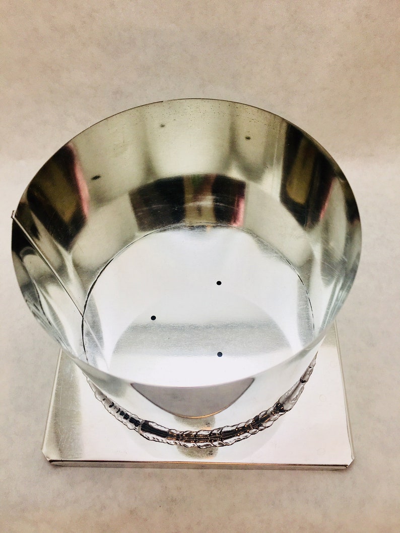 May include: A silver metal candle mold with a round top and a square base. The mold has three small holes in the bottom.