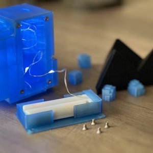 Puede incluir: Un cubo de pl&aacute;stico azul con una tapa de pl&aacute;stico transparente y una cadena de luces blancas en su interior. El cubo est&aacute; desmontado y la tapa est&aacute; quitada. Hay cuatro peque&ntilde;as piezas de pl&aacute;stico azul y una pieza triangular negra sobre una superficie de madera.