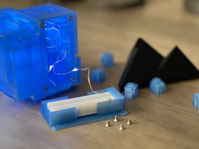 Puede incluir: Un cubo de pl&aacute;stico azul con una tapa de pl&aacute;stico transparente y una cadena de luces blancas en su interior. El cubo est&aacute; desmontado y la tapa est&aacute; quitada. Hay cuatro peque&ntilde;as piezas de pl&aacute;stico azul y una pieza triangular negra sobre una superficie de madera.