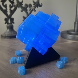 Puede incluir: Un objeto azul en forma de cubo impreso en 3D sobre un soporte triangular negro. El cubo est&aacute; iluminado desde el interior, lo que le da un brillo azul. Hay varios cubos azules m&aacute;s peque&ntilde;os dispersos alrededor de la base.